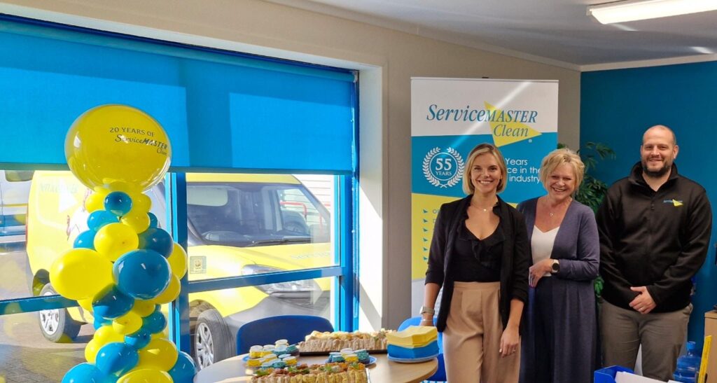 Three people stand next to a table with food on it, next to a yellow and blue balloon arch